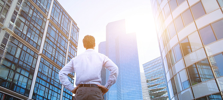 dow_39921096418-submkthero-man-skyscrapers-743x335 呈现一名商人的背影,此刻他站在玻璃摩天大楼之间,向上凝视着大楼。